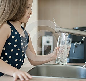 Portrait of a little caucasian girl gaining a glass of tap clean water. Kitchen faucet. Cute curly kid pouring fresh water