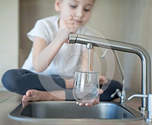 Portrait of little caucasian girl gaining a glass of tap clean water. Kitchen faucet. Kid pouring fresh water from filter tap.