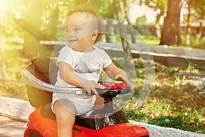 Portrait of a little cheerful baby in white shirt sitting half turned back on red push car in park or garden in sun rays