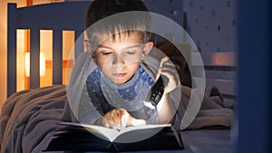 Portrait of little concentrated boy lying in bed and reading book with flashlight