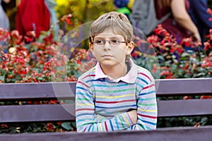 Portrait of little cool kid boy with eye glasses sitting on bank in park. Happy healthy child having fun on warm sunny