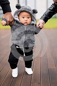 Portrait of little cute baby boy making first steps, holding hands of father and mother, looking at the camera