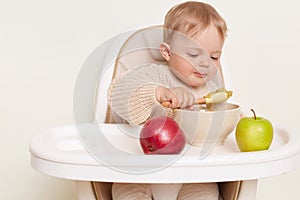 Portrait of little cute charming baby eating food from a plate with spoon against while background, sitting with apples around,