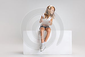 Portrait of little cute girl in casual clothes sitting on big box isolated on white studio background. Happy childhood