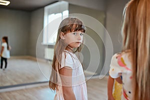 Portrait of a little cute girl looking at camera while standing against mirror in the dance studio. Group of children