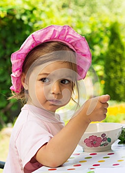 Portrait of little cute girl in a purpule cap eats with appetite a breakfast from a plate with the drawn flower in outdoor cafe