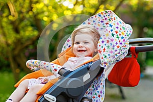 Portrait of little cute smiling toddler girl sitting in stroller or pram and going for a walk. Happy cute baby child