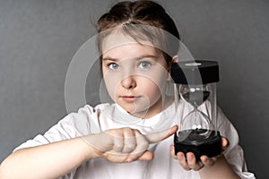 Portrait of a little cute teenage girl holding an hourglass while pointing at it with her index finger.