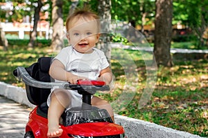 Portrait of a little driver: happy infant child with surprised face sitting barefoot on a red push car outdoor in the park