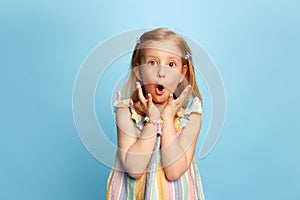 Portrait of little emotive girl, child standing with shocked face, looking at camera against blue studio background