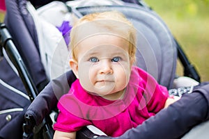Portrait of a little funny child girl blond with blue eyes sitting in a baby stroller in the summer for greens. Trinasport for a c