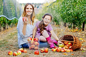 Portrait of little girl and beautiful mother with red apples in organic orchard. Happy woman and kid daughter picking