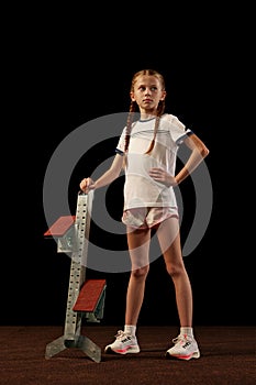 Portrait of little girl, beginner athlete in sportswear posing with starting blocks isolated over dark background, Sport