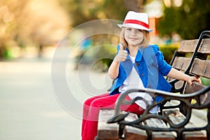 Portrait of little girl on bench in a park showing thumb up
