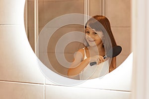 Portrait of little girl combing her hair in the bathroom, doing morning beauty procedures by her own while standing in front of