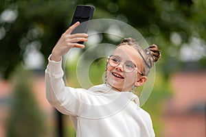 Portrait of a little girl in glasses with a smartphone taking a selfie in summer park