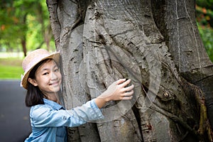 Portrait of little girl in hat with smiling,hugging large tree trunk with arms around tree and looking at camera at outdoor park,