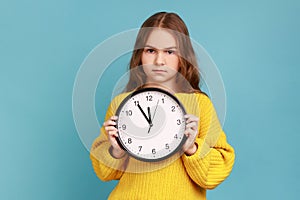 Portrait of little girl holding big wall clock in hands, looking at camera with serious expression.