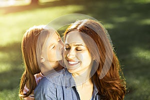 Portrait of little girl kissing her mother in the park with blurred background