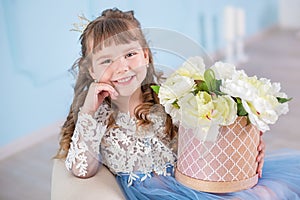 Portrait of little girl in luxurious dress posing in decorated blue studio with bouquet of flowers on retro sofa divan