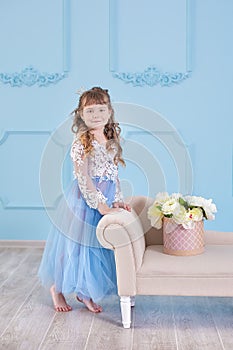 Portrait of little girl in luxurious dress posing in decorated blue studio with bouquet of flowers on retro sofa divan