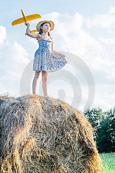 Portrait of little girl playing, standing on haystack in field. Playing flying toy airplane.Light sunny day. Low angle