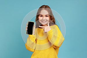 Portrait of little girl pointing finger at blank screen of mobile phone, looking smiling to camera.