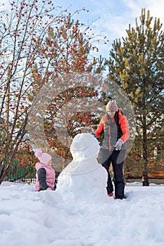 Portrait of little girl sitting on snow and mother making snowman on backyard in evening with rowan and fir trees in