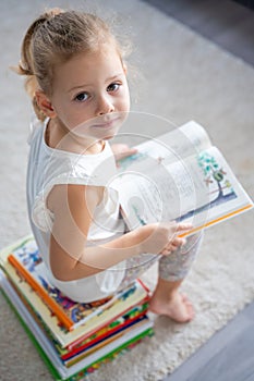 Portrait of Little girl is sitting on stack of children& x27;s books and leafing through a book with fairy tales