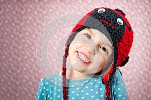 Portrait of little girl smiling and being happy. Child is wearing a woolen cap in the shape of ladybird on a background with red