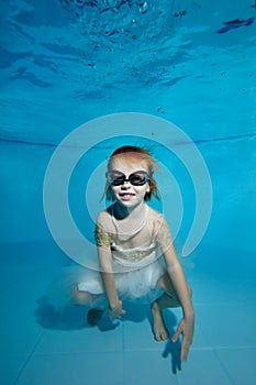 Portrait of a little girl under water at the bottom of the pool. She smiles and looks at the camera. Baby