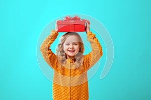 Portrait of a little girl in a warm mustard knitted sweater on a cyan background. A child holds a red box with a gift on the head.