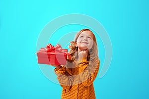 Portrait of a little girl in a warm mustard knitted sweater on a cyan background. The child looks dreamily up and holds a red gift