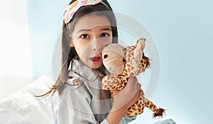 Portrait of a little girl wearing pajama in stripes blue  and white, smiling broadly, playing with toy giraffe in the bed,