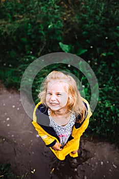 Portrait of little girl wearing summer dress, yellow, blue windbreaker, winking, standing on wet pavement after rain.