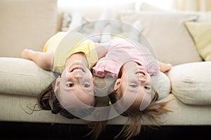 Portrait of little girls laughing lying upside down. Cheerful kids sisters having fun on sofa at home