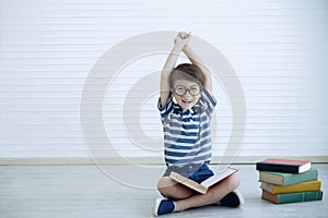 Portrait of little happy Caucasian boy wear glasses sitting on the floor looking at camera and reading a big book at home.