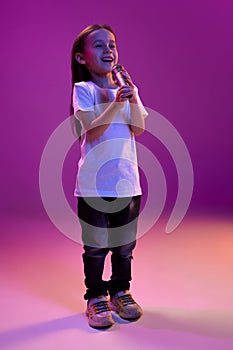 Portrait of little kid, child in white T-shirt posing, cheerfully singing in microphone over purple background in neon