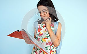 Portrait of a little kid girl 7-8 years old dressed in colorful dress, wearing eyeglasses read a book  on light blue