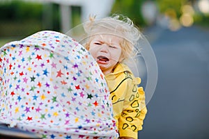 Portrait of little sad toddler girl sitting in stroller and going for a walk. Crying baby child does not want sitting in