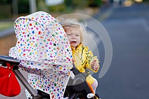 Portrait of little sad toddler girl sitting in stroller and going for a walk. Crying baby child does not want sitting in
