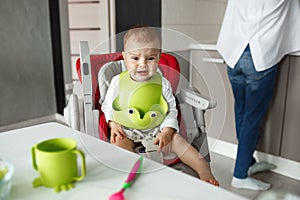Portrait of little scared baby boy sitting in baby chair in kitchen, crying and screaming while mother cooking him food.