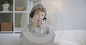 Portrait of little schooler boy at home, chatting online by notebook with internet connection, sitting at living room at