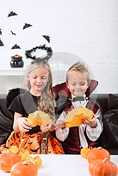 portrait of little siblings in halloween costumes sitting on sofa at table with pumpkins
