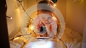 Portrait of little smart boy reading big story book at night. Child playing in toy cardboard house. Concept of child
