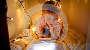 Portrait of little smart boy reading big story book at night. Child playing in toy cardboard house. Concept of child