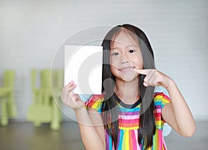 Portrait little smiling Asian child girl holding blank white paper card in her hand with pointing. Kid showing empty paper note