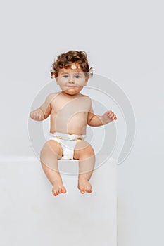 Portrait of little sweet toddler boy, baby in diaper calmly sitting isolated over white studio background