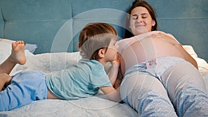 Portrait of little toddler boy in pajamas lying with pregnant mother in bed and talking to unborn baby in big belly