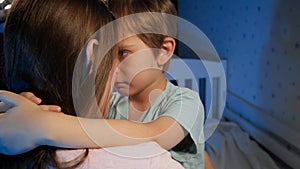 Portrait of little upset and sad toddler boy hugging and kissing his mother before going to sleep at night. Loving and